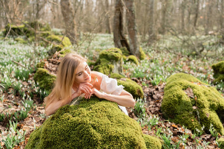 Snowdrops galanthus blond. A girl in a white dress lay down on a stone in the moss in a meadow with snowdrops in a spring forestの写真素材
