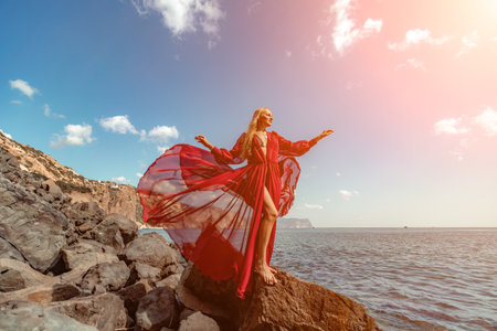 Red dress sea woman. A blonde with flowing hair in a long red dress sits on a rock near the sea. The concept of trips, a photo shoot at the seaの写真素材