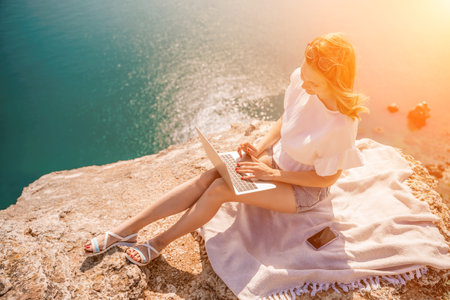 Freelance woman working on a laptop by the sea, typing away on the keyboard while enjoying the beautiful view, highlighting the idea of remote work.の写真素材