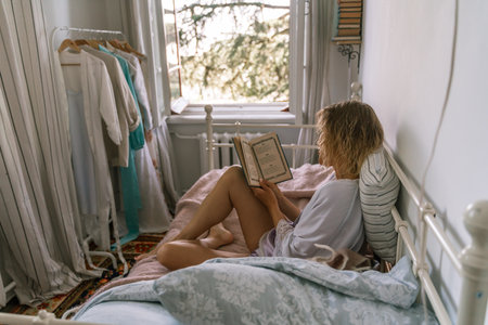 Woman reading a book in bed, lying on her stomach smiling happy and relaxed on a leisure day at homeの写真素材