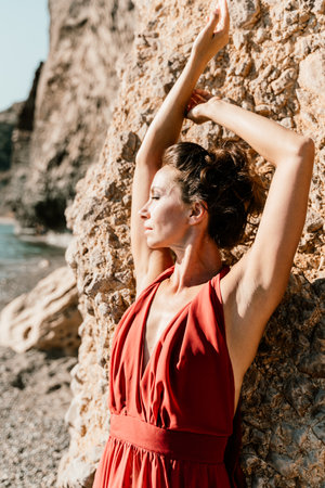 Woman red dress sea. Woman in a long red dress posing on a beach with rocks on sunny day. Girl on the nature on blue sky background.の写真素材