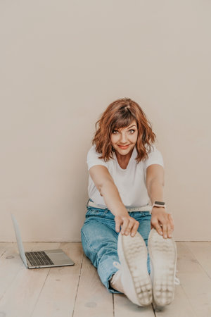 A brunette sits on the floor with a laptop on a beige wall background. She is wearing a white T-shirt, jeans and white sneakers.の写真素材