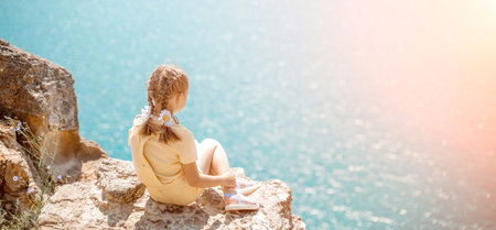 Happy girl perched atop a high rock above the sea, wearing a yellow jumpsuit and braided hair, signifying the concept of summer vacation at the beach.の写真素材