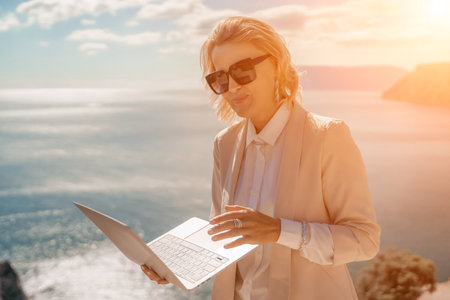 Freelance women sea. She is working on the computer. Good looking middle aged woman typing on a laptop keyboard outdoors with a beautiful sea view. The concept of remote work.の写真素材