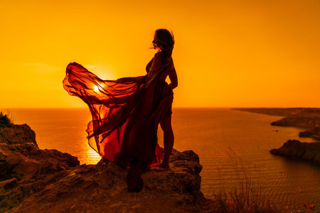 Woman sunset sea red dress, side view a happy beautiful sensual woman in a red long dress posing on a rock high above the sea on sunset.の写真素材