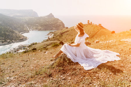 Happy woman in a white dress and hat stands on a rocky cliff above the sea, with the beautiful silhouette of hills in thick fog in the background.の写真素材