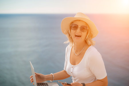 Freelance women sea working on the computer. Good looking middle aged woman typing on a laptop keyboard outdoors with a beautiful sea view. The concept of remote work.の写真素材