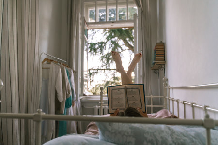 Woman reading a book in bed, lying on her stomach smiling happy and relaxed on a leisure day at homeの写真素材