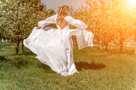 Woman white dress park. A woman in a white dress runs through a blossoming cherry orchard. The long dress flies to the sides, the bride runs rejoicing in life.の写真素材