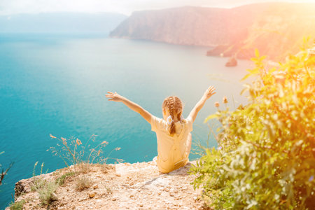 Happy girl perched atop a high rock above the sea, wearing a yellow jumpsuit and braided hair, signifying the concept of summer vacation at the beach.の写真素材