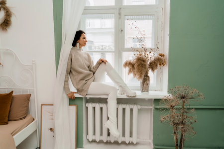 Free time for rest. Profile of a beautiful woman sitting on a white window sill at home, holding a cotton plant in her hands. She is wearing a sweater, leggings and white boots.の写真素材