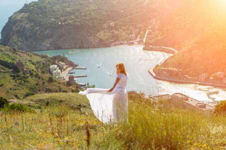 Happy woman in a white dress and hat stands on a rocky cliff above the sea, with the beautiful silhouette of hills in thick fog in the background.の写真素材