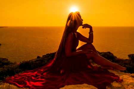 Woman sunset sea red dress, side view a happy beautiful sensual woman in a red long dress posing on a rock high above the sea on sunset.の写真素材