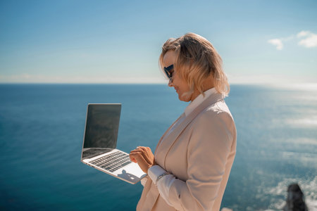 Freelance women sea working on the computer. Good looking middle aged woman typing on a laptop keyboard outdoors with a beautiful sea view. The concept of remote work.の写真素材