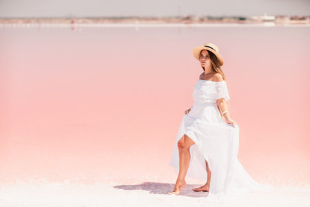 Woman in pink salt lake. She in a white dress and hat enjoys the scenic view of a pink salt lake as she walks along the white, salty shore, creating a lasting memory.の写真素材