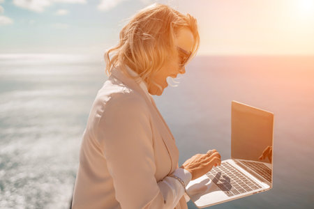 Freelance women sea working on the computer. Good looking middle aged woman typing on a laptop keyboard outdoors with a beautiful sea view. The concept of remote work.の写真素材