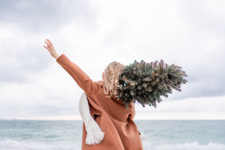 Blond woman Christmas tree sea. Christmas portrait of a happy woman walking along the beach and holding a Christmas tree on her shoulder. She is wearing a brown coat and a white suit.の写真素材