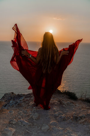 Woman sunset sea red dress, back view a happy beautiful sensual woman in a red long dress posing on a rock high above the sea on sunset.の写真素材