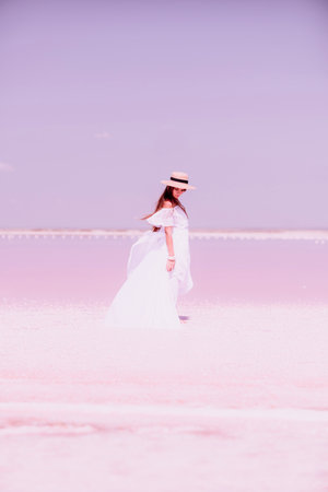 Woman in pink salt lake. She in a white dress and hat enjoys the scenic view of a pink salt lake as she walks along the white, salty shore, creating a lasting memory.の写真素材