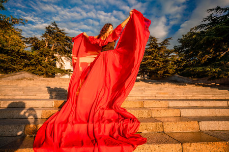Sunrise red dress. A woman in a long red dress against the backdrop of sunrise, bright golden light of the suns rays. The concept of femininity, harmony.の写真素材