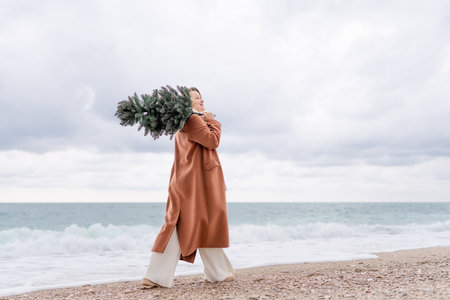 Blond woman Christmas tree sea. Christmas portrait of a happy woman walking along the beach and holding a Christmas tree on her shoulder. She is wearing a brown coat and a white suit.の写真素材