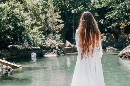 a beautiful woman in a long white dress looks into the distance at a beautiful lake with swans rear viewの写真素材