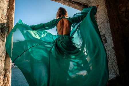 Rear view of a happy blonde woman in a long mint dress posing against the backdrop of the sea in an old building with columns. Girl in nature against the blue sky.の写真素材