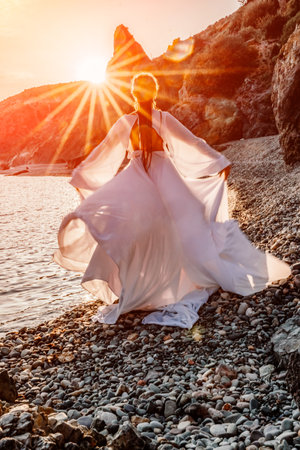 Mysterious woman silhouette long hair walks on the beach ocean water, sea nymph wind listens to the wave. Throws up a long white dress, a divine sunset. Artistic photo from the back without a faceの写真素材