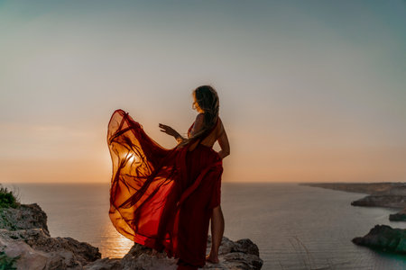 Woman sunset sea red dress, side view a happy beautiful sensual woman in a red long dress posing on a rock high above the sea on sunset.の写真素材
