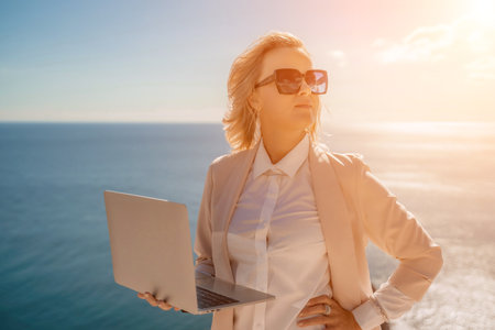 Freelance women sea. She is working on the computer. Good looking middle aged woman typing on a laptop keyboard outdoors with a beautiful sea view. The concept of remote work.の写真素材