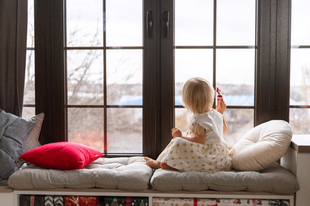 Girl 2 years old sits on the windowsill. A little blonde girl in a white dress on the windowsill looks out the window and holds a candy in her hand.の写真素材
