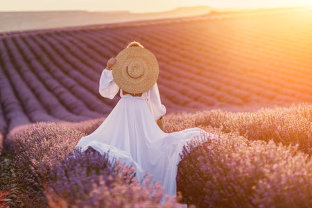 Happy woman in a white dress and straw hat strolling through a lavender field at sunrise, taking in the tranquil atmosphere.の写真素材
