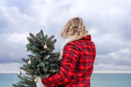 Blond woman holding Christmas tree by the sea. Christmas portrait of a happy woman walking along the beach and holding a Christmas tree in her hands. Dressed in a red coat, white suit.の写真素材