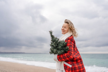 Blond woman holding Christmas tree by the sea. Christmas portrait of a happy woman walking along the beach and holding a Christmas tree in her hands. Dressed in a red coat, white dress.の写真素材