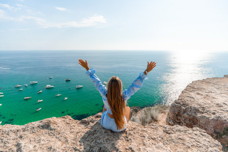 Woman travel sea. Happy woman in a beautiful location poses on a cliff high above the sea, with emerald waters and yachts in the background, while sharing her travel experiencesの写真素材