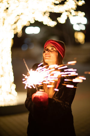 Woman holding sparkler night while celebrating Christmas outside. Dressed in a fur coat and a red headband. Blurred christmas decorations in the background. Selective focusの写真素材