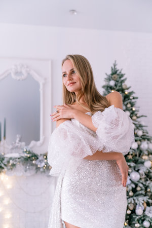 The blonde in the Christmas room. A beautiful blonde woman in a shiny light short dress with a train stands in a beautiful bright room decorated with a festive interior with a Christmas tree.の写真素材