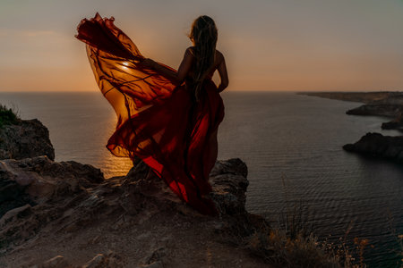 Woman sunset sea red dress, side view a happy beautiful sensual woman in a red long dress posing on a rock high above the sea on sunset.の写真素材