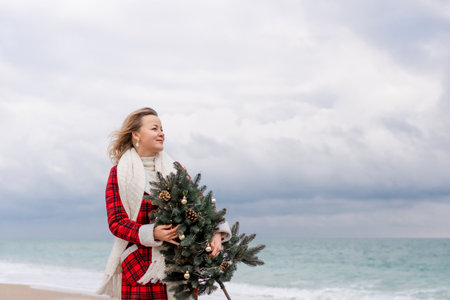 Blond woman holding Christmas tree by the sea. Christmas portrait of a happy woman walking along the beach and holding a Christmas tree in her hands. Dressed in a red coat, white dress.の写真素材