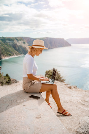 Freelance women sea working on the computer. Good looking middle aged woman typing on a laptop keyboard outdoors with a beautiful sea view. The concept of remote work.の写真素材