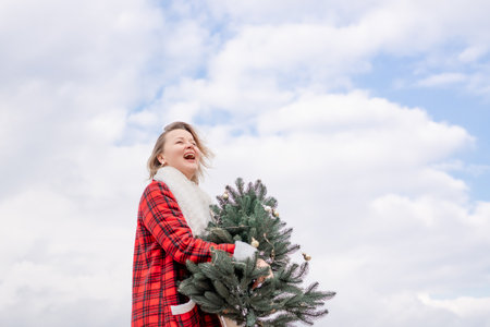Blond woman holding Christmas tree by the sea. Christmas portrait of a happy woman walking along the beach and holding a Christmas tree in her hands. Dressed in a red coat, white suit.の写真素材