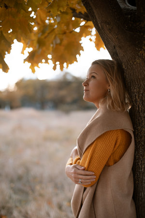 The blonde stands near the autumn tree. Thoughtful woman looks ahead, dressed in a yellow dress. Autumn contentの写真素材