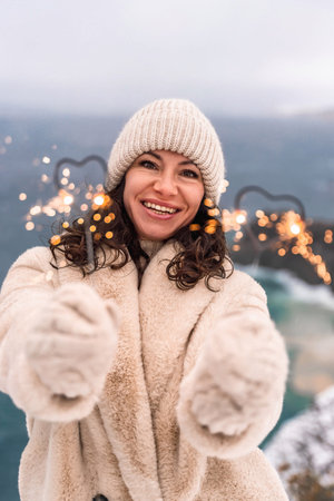 Outdoor winter portrait of happy smiling woman, light faux fur coat holding heart sparkler, posing...
