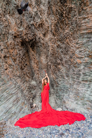 woman sea red dress. Woman with long hair on a sunny seashore in a red flowing dress, back view, silk fabric waving in the wind. Against the backdrop of the blue sky and mountains on the seashore.の写真素材