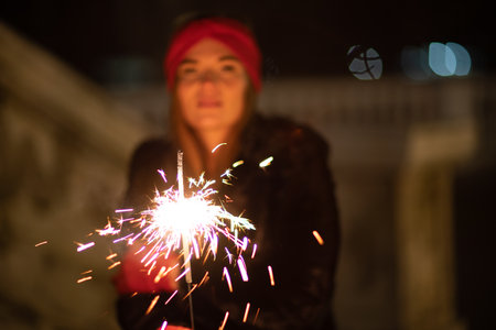 Woman holding sparkler night while celebrating Christmas outside. Dressed in a fur coat and a red headband. Blurred christmas decorations in the background. Selective focusの写真素材