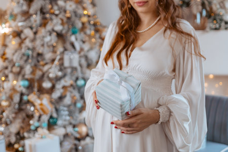 Pregnant woman holds a gift on her stomach against Christmas tree with lights. family holiday concept Christmas holidays.の写真素材