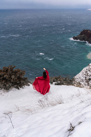 Woman red dress snow sea. Happy woman in a red dress in the snowy mountains by the emerald sea. The wind blows her clothes, posing against sea and snow background.の写真素材