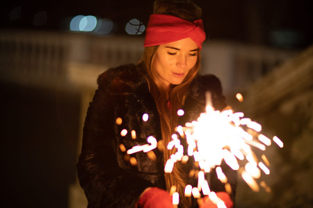 Woman holding sparkler night while celebrating Christmas outside. Dressed in a fur coat and a red...