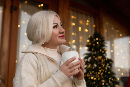 Happy blonde at home at Christmas time, drinking hot beverage from the white mug. Christmas tree in the background. Celebrating Christmas and New Year at home in cozyの写真素材