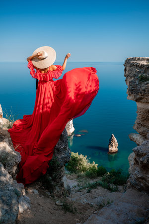 Red dress sea woman. Happy woman with flowing hair in a long flowing red dress stands on a rock near the sea. Travel concept, photo session at seaの写真素材